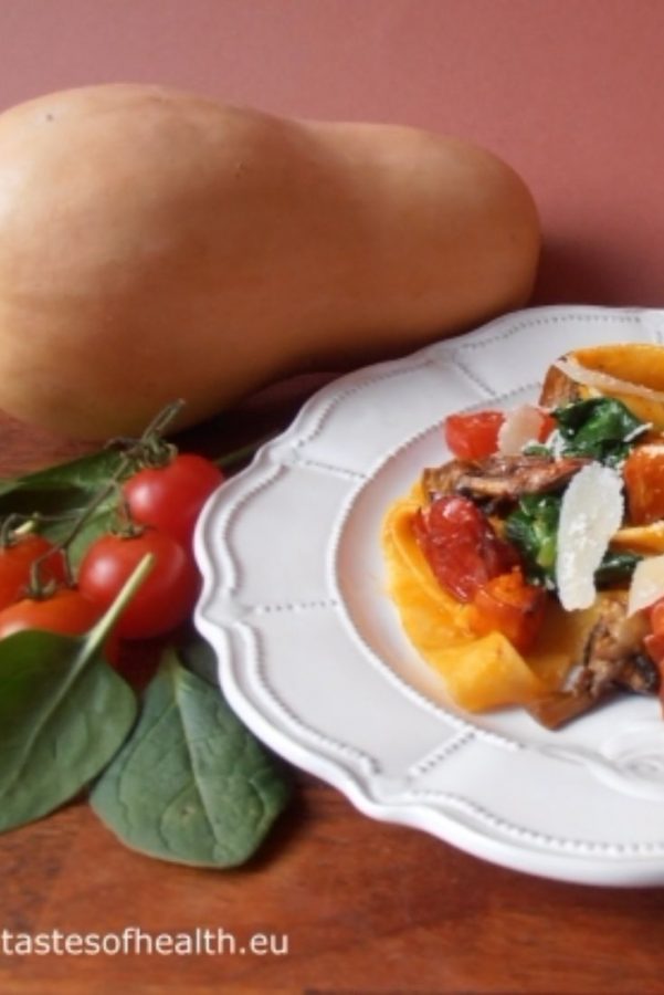 An image showing 2 Method Pasta with Squash, Mushrooms and Spinach on a white patterned plate on a wooden surface, surrounded by a whole squash, spinach, cherry tomatoes, and mushrooms, with a purple background.