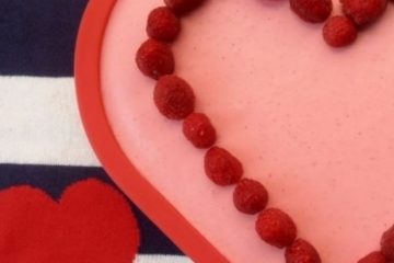 A heart-shaped cake decorated with raspberries on a white and blue background, with two red heart next to it.