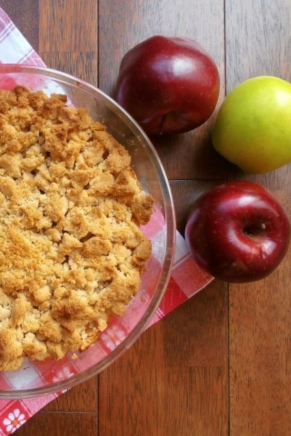 An image of an apple crumble in a transparent bowl on a checkered napkin, surrounded by two red apples and one green apple, on a wooden floor.