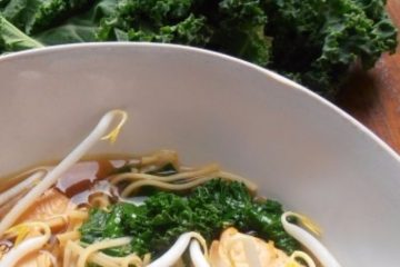 An image showing Asian Soup with Salmon and Kale with bean sprouts in a white bowl with kale next to it on a wooden surface.