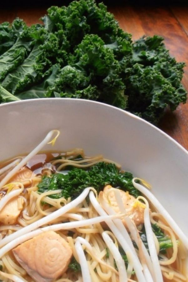 An image showing Asian Soup with Salmon and Kale with bean sprouts in a white bowl with kale next to it on a wooden surface.