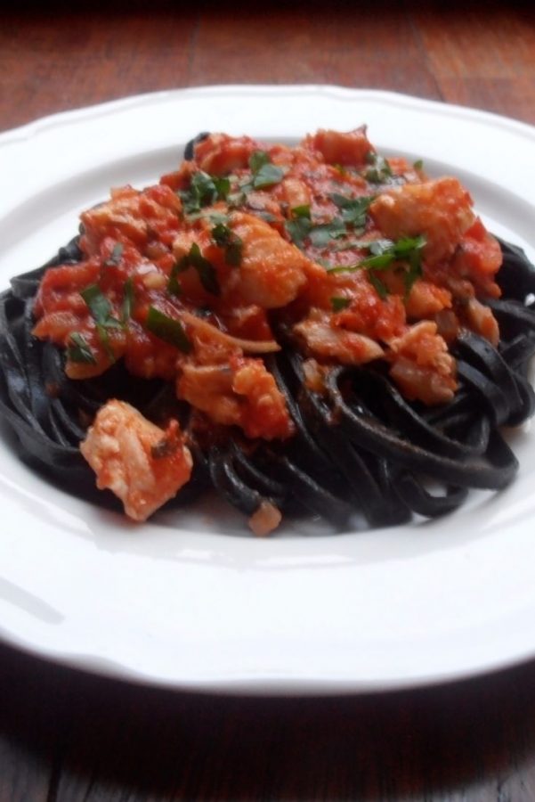 An image showing Black Pasta with Cod and Tomato Sauce on a white plate on a wooden surface.