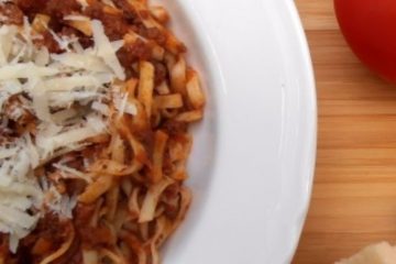 An image showing half a white bowl of traditional bolognese sauce with tagliatelle, topped with parmesan cheese, on a wooden board with a tomato and some parmesan next to the bowl.