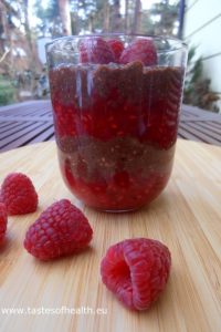 An image of a glass with layers of chocolate chia pudding and raspberry mousse with a few raspberries next to it.