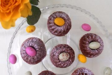 An image showing Chocolate Cupcakes, with mini eggs in the center of them, on a glass plate, with more mini eggs on the plate, yellow roses next to the plate, and a white tablecloth.