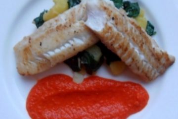 An image showing cod with spinach, potatoes, and a red pepper sauce below, on a white plate on a wooden floor.