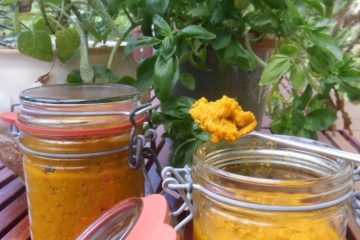 An image showing two jars of Concentrated Vegetable Stock Paste, with one jar open and with a teaspoon full of the paste on top of the jar- both jars are on a wooden table and have greenery behind them.