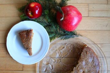 An image of Gingerbread Spiced Apple Cake on a glass plate. There is also a slice of the cake on a white plate, a red apple, a pomegranate on a green brunch next to it.