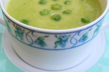 An image showing Green Pea Soup in a white cup with a blue flower pattern on the edge, on a white plate with blue on the edge, on a wooden surface.