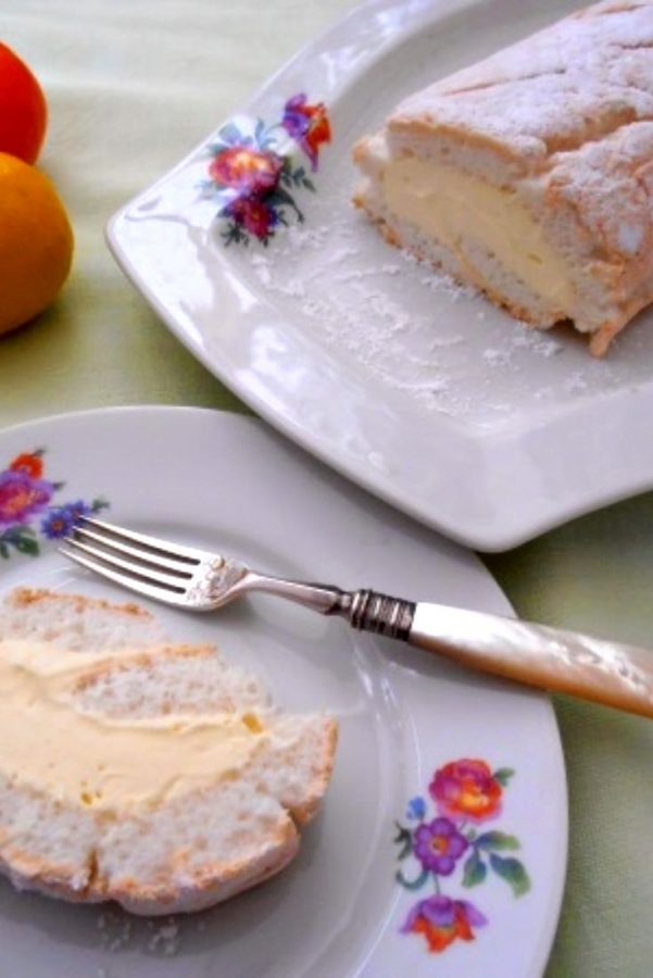 An image showing Lemon Meringue Roulade on a long white plate with flower decorations along the side, next to a white plate with a piece of cake and a fork on it, on a green tablecloth.