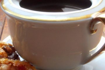 An image showing a teacup with coffee in it, on a plate, with chocolate florentines: one on top of the teacup and another on the plate, on a wooden floor.