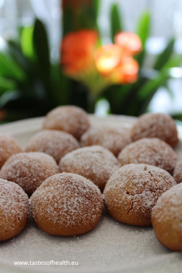 An image of golden-brown Marzipan Cookies, covered in powdered sugar, on a white plate. There is a blooming plant in the background.