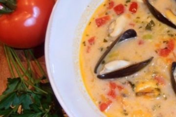 An image showing Mussel Soup with Vegetables in a white plate next to a green vegetable and a tomato on a wooden surface.