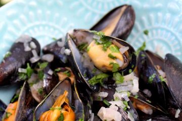 An image of Mussels in White Wine Sauce on a blue plate. There are pink blooms in the background. Learn how to clean mussels.