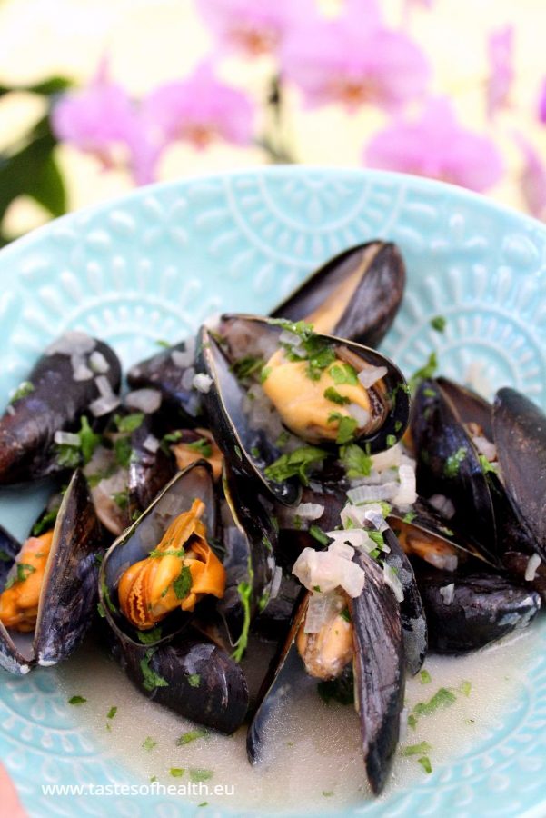 An image of Mussels in White Wine Sauce on a blue plate. There are pink blooms in the background. Learn how to clean mussels.