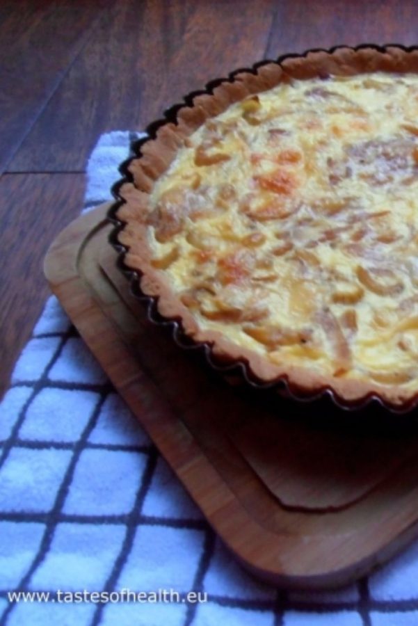 An image showing an Onion Tart with Gluten-Free Pastry Crust in a black plate, on a wooden board, on a black-and-white striped towel, on a wooden floor.