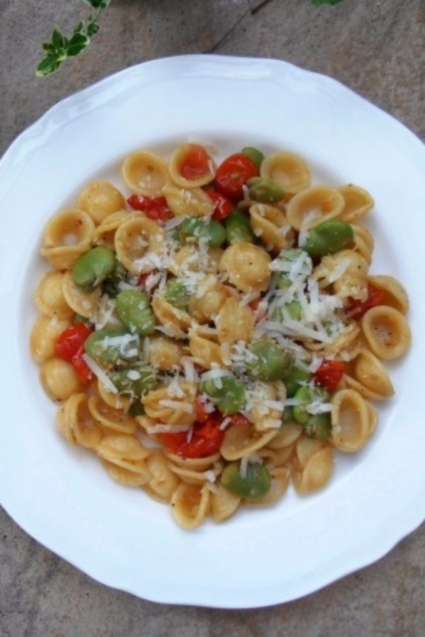 An image showing Orecchiette with Broad Beans and Tomatoes, topped with parmesan, in a white bowl, on a marble surface.