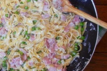 An image of mascarpone pasta with ham and green peas in a black bowl, with a wooden spoon also in the bowl, on top of a green towel, which is on a wooden floor.