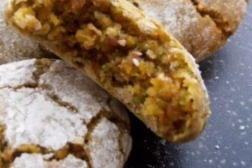 An image of pistachio cookies dusted with icing sugar on a black board. The inside of one of the biscuits is visible.