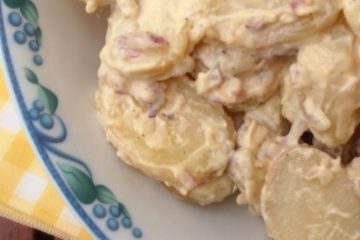 An image of a Potato Salad in a blue and white floral-patterned bowl, on top of a yellow and white checkered place mat and a wooden table.