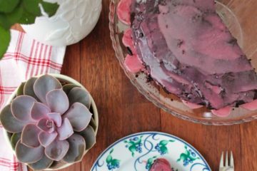 An image of Quinoa Chocolate Cake set on a glass plate and a slice of the quinoa cake on a white plate with green and blue patterns. There are pink roses in a white vase, a plant and a white and red checkered cloth next to the dishes.