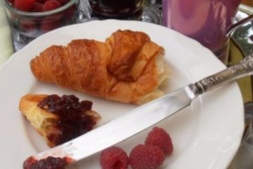 An image of a silver tray with a jar of Raspberry Jam, a mug of coffee, a bowl of raspberries, and a white plate with a croissant with raspberry jam.