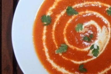 An image showing Roasted Tomato Soup with a white swirl on top, in a white bowl on a wooden surface.