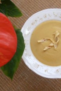 An image of a spicy parsnip soup in a white plate, on a wicker table, with a red plant with green leaves next to it.