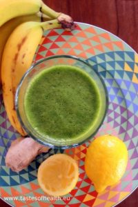 An image of a Spinach, Ginger and Banana Smoothie in a glass on a colorful plate, with a banana, ginger and lemon next to it.