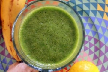 An image of a Spinach, Ginger and Banana Smoothie in a glass on a colorful plate, with a banana, ginger and lemon next to it.