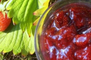 An image of a glass container with strawberry jam standing on grass with fresh strawberry fruit and leaves next to it.
