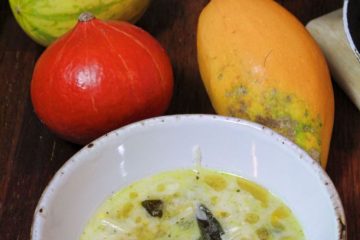 An image of Sweetcorn and Spaghetti Squash Soup ina white bowl. There are 3 different types of pumkin next to it as well as a pink rose in a glass vase.