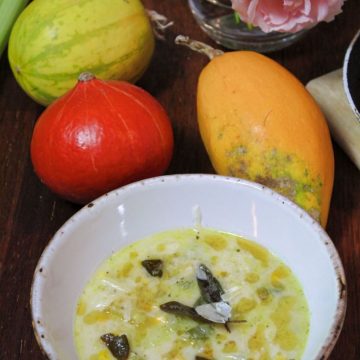 An image of Sweetcorn and Spaghetti Squash Soup ina white bowl. There are 3 different types of pumkin next to it as well as a pink rose in a glass vase.