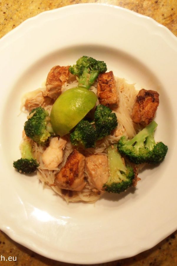 An image showing Teriyaki Salmon with Broccoli and Rice Vermicelli with a lime on top, on a white bowl.