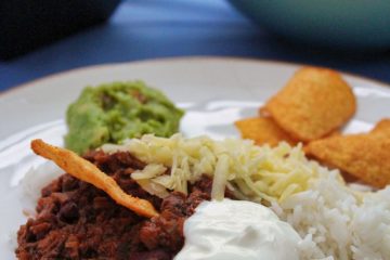 Vegetarian Chilli Con Carne served on a white plate with rice, grated cheese, guacamole and tortilla chips.