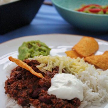 Vegetarian Chilli Con Carne served on a white plate with rice, grated cheese, guacamole and tortilla chips.