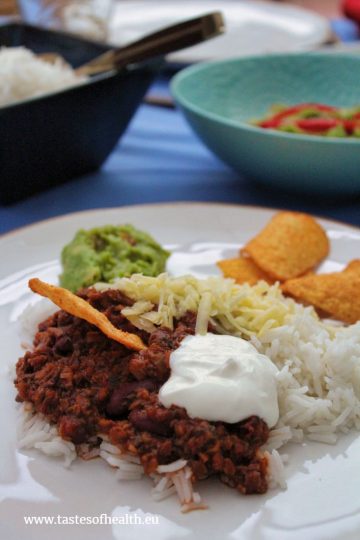 Vegetarian Chilli Con Carne served on a white plate with rice, grated cheese, guacamole and tortilla chips.