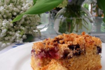 An image of a slice of an almond berry cake on a white plate with a vase and flowers in the background.