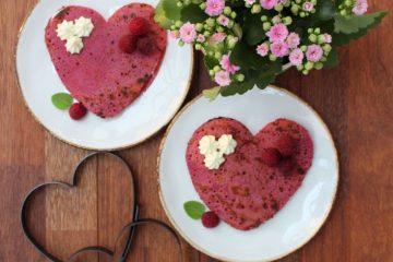 Two heart-shaped pink beet pancakes on white plates with a pink blloming plant and 2 metal hearts next to them.