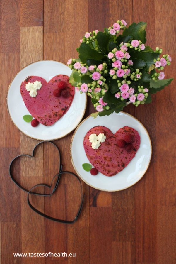 Two heart-shaped pink beet pancakes on white plates with a pink blloming plant and 2 metal hearts next to them.