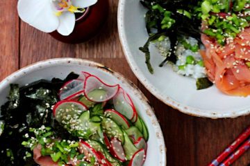 An image of sushi bowl x2 with smoked salmon, shredded nori, slices of cucumber and radish on top. There are black and red chopsticks as well as a white bloom next to the bowls
