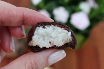 An image of hand held, half eaten Coconut Bounty Balls. Other balls are on a golden plate in the background. There is also a pot of light pink carnations there.