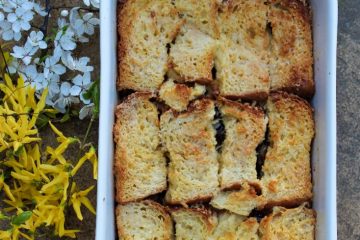 An image of Chocolate Bread and Butter Pudding in a white dish. There are white and yellow blooms next to the dish.