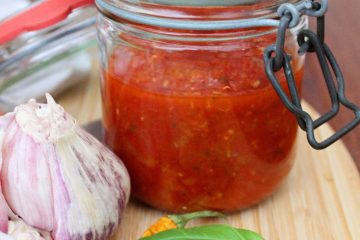 An image of a jar with Homemade Tomato Sauce. There are garlic cloves, basil leaf and yellow pepper next to the jar.