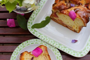 An image of a Rhubarb Cake: a piece and the rest of the roll. Both are on white plates. There is also a vase with colorful blooms next to them.