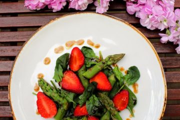 An image of Spinach Salad with Strawberries and Asparagus on a white plate. The plate is on a brown wooden table and there are pink blooms next to it.