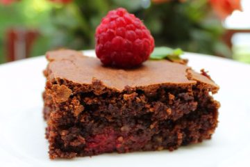 An image of Gluten Free Brownies on a white plate with a raspberry on top. There are peach-coloured roses in the background.
