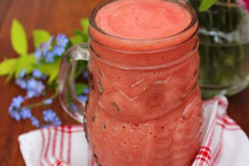 An image of Strawberry and Pineapple Smoothie in a glass on a white and red teatowel. There are blue and pink flowers in the background.
