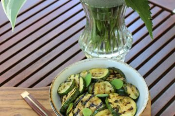 An image of Courgette Salad in a white bowl standing on a wooden board. The board is on a table and there is a vase with colourful flowers next to it.