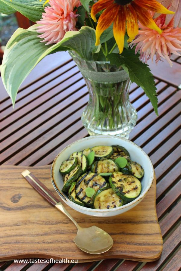 An image of Courgette Salad in a white bowl standing on a wooden board. The board is on a table and there is a vase with colourful flowers next to it.